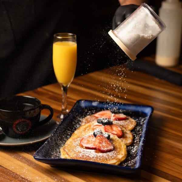 Powdered sugar being sprinkled over pancakes with berries, a mimosa, and coffee on a wooden table.