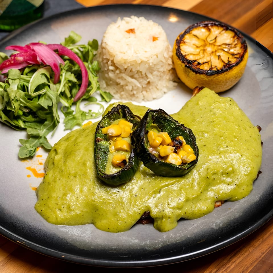 A vibrant green sauce dish with grilled peppers, corn, rice, arugula, pickled onions, and a charred lemon.