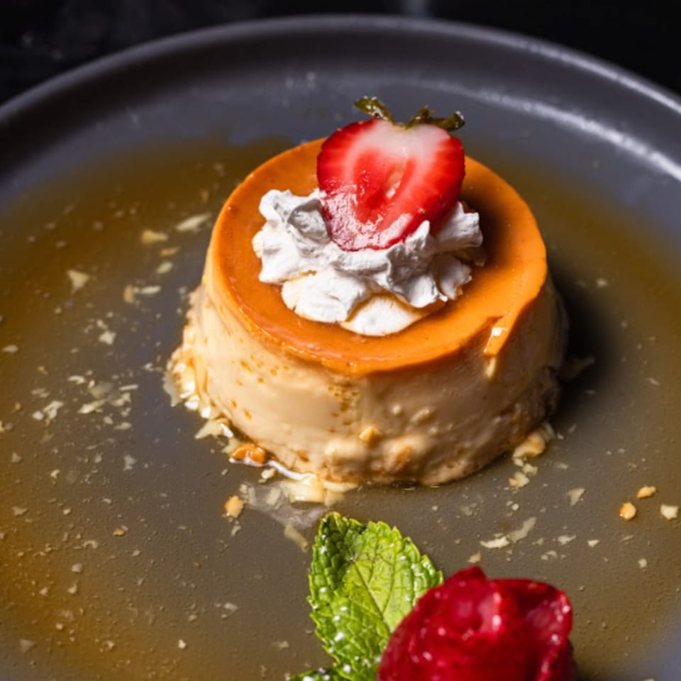 Flan dessert with caramel sauce, whipped cream, a strawberry half, and mint on a gray plate.