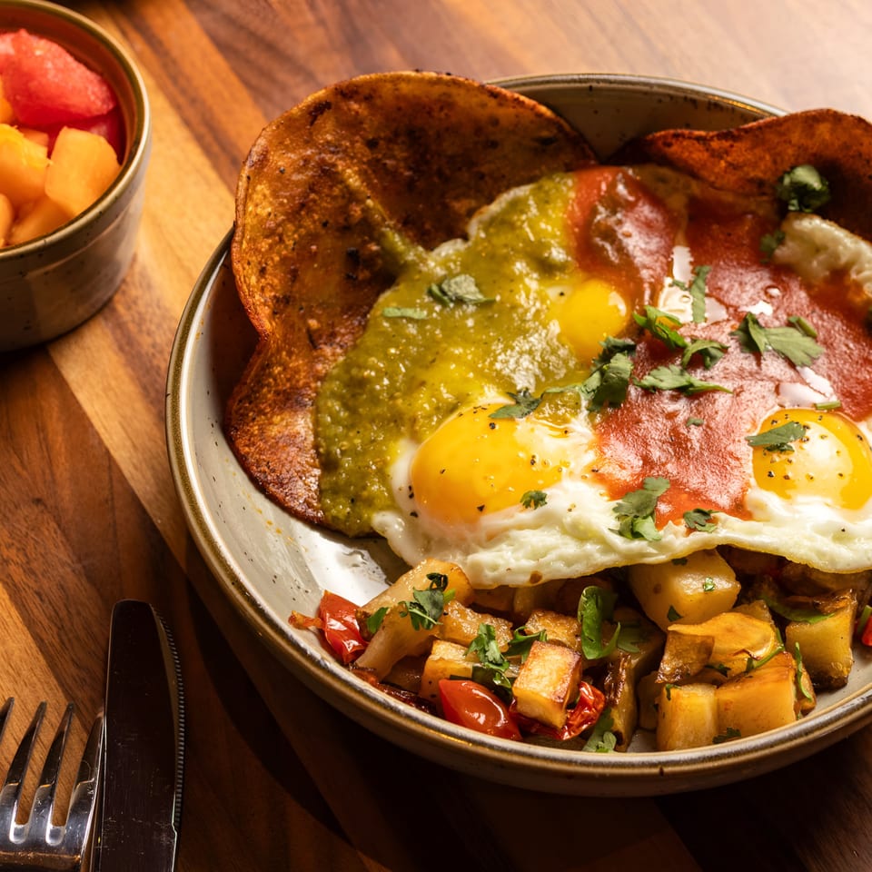 Huevos rancheros with green and red salsa, fried eggs, potatoes, and a side of fruit.