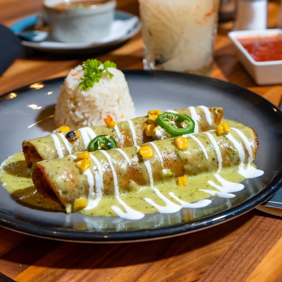 Two enchiladas covered in green sauce, sour cream, corn, and jalapeños, served with rice.